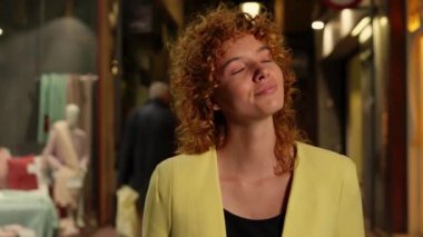 Genç bir kadın gülümsüyor ve etrafa bakıyor. Canlı bir şehir caddesinde, vitrinlerle dolu bir gece yürürken.