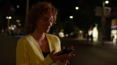 Kıvırcık kızıl saçlı genç bir kadın gece bir caddede akıllı telefonunu kullanırken gülümsüyor.