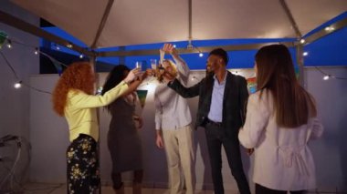 Çok ırklı genç yetişkinler kadeh kaldırıyor ve gecenin bir yarısında çatıda kutlama yapıyor, içkinin tadını çıkarıyor ve birlikte neşeli anılar yaratıyorlar.