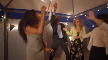 Çok ırklı arkadaşlar, yaz geceleri canlı bir çatı partisinde dans ederken ve havai fişek tutarken sevinçle kutlama yapıyorlar.