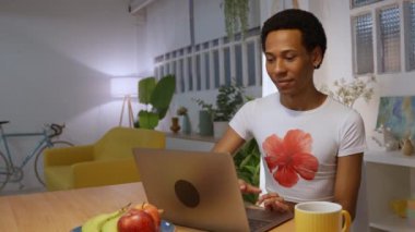 Genç yetişkin bir adam mutfak tezgahında otururken dizüstü bilgisayarından bir şeye olumlu tepki veriyor.