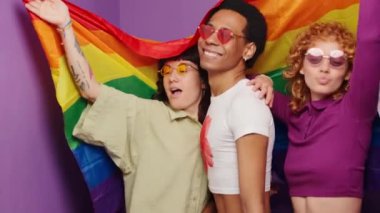 Üç arkadaş bir fotoğraf kabininde çekilen bir dizi fotoğrafta gökkuşağı bayraklarıyla gülümsüyor ve poz veriyorlar.