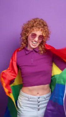 Kızıl saçlı lgbtq eylemcisi gökkuşağı bayrağı sallıyor, gururu ve özgürlüğü mor arka planda kutluyor, yavaş çekim