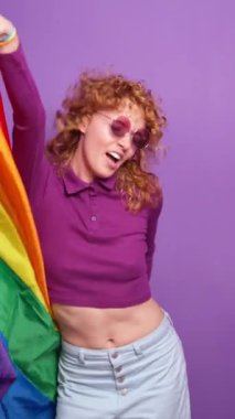 Gökkuşağı bayrağıyla dans eden enerjik kızıl saçlı kadın mor arka planda lgbtq gururunu kutluyor.