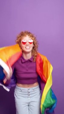 Kızıl saçlı lgbtq aktivisti kalp şeklinde güneş gözlüklü gökkuşağı bayrağıyla dans eden mor arka planda gurur ve çeşitliliği kutlayan, ağır çekim