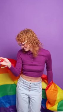 Kızıl saçlı kadın Igbt gökkuşağı bayrağıyla canlı mor bir arka planda dans ederek aşkı ve çeşitliliği kutluyor.