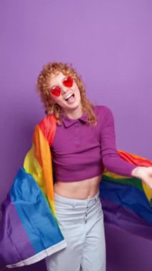 Kızıl saçlı lgbtq aktivisti, kalp şeklinde güneş gözlüklü dans eden ve parlak mor arka planda gökkuşağı bayrağı sallayan.