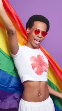 Kırmızı kalp şeklindeki güneş gözlüklü genç adam mor arka planda gökkuşağı bayrağıyla dans ediyor, ağır çekimde lgbtq gururunu kutluyor.