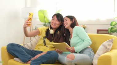 İki neşeli genç kadın sarı bir kanepede oturup, akıllı telefonlarıyla selfie çekiyorlar ve tablet okuyup, birlikte vakit geçiriyorlar.