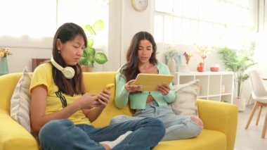 İki arkadaş sarı bir kanepede birlikte vakit geçiriyorlar, cep telefonu ve tablet arasındaki içeriği paylaşıyorlar.