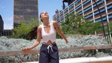 Genç kadın enerjik dans figürlerini canlı bir şehir parkında sergiliyor. Yeşillik ve modern mimari, canlı atmosferi geliştirir.