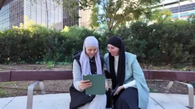 İki Müslüman iş kadını, modern bir şehir parkında dijital stratejileri tartışan bir tablet üzerinde işbirliği yapıyorlar.