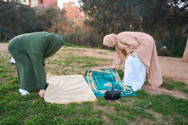 Two muslim women wearing traditional clothing are laying down prayer rugs on grass in a park, preparing for prayer clipart