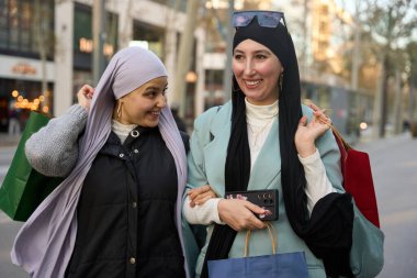 Başörtüsü takan iki mutlu Müslüman kadın, ellerinde alışveriş torbalarıyla şehir caddesinde kol kola yürüyorlar ve günlerinin tadını çıkarıyorlar.
