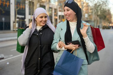 Başörtüsü takan iki gülümseyen Müslüman kadın, ellerinde alışveriş çantaları ve akıllı bir telefonla, şehir caddesinde kol kola yürüyorlar.