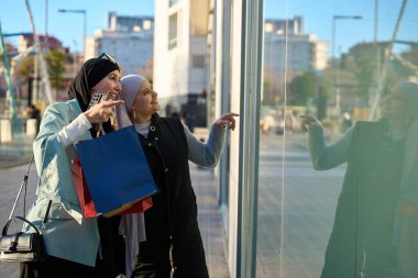 Alışveriş torbaları taşıyan, alışveriş çılgınlığından zevk alan, vitrindeki eşyaları işaret eden iki neşeli Müslüman kadın.