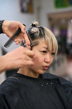 Kuaför tarak ve makas kullanıyor, kuaförde kısa sarı saçlı genç bir kadın modelin saçını kesiyor.