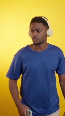 Hevesli genç adam müziği, dansı ve neşeli kulaklıkları ve parlak sarı arka planda akıllı telefonuyla ifade etmeyi seviyor.