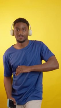 Kulaklık takan, akıllı telefon tutan, dans eden ve sarı arka planda izole edilmiş müzik dinleyen mutlu genç adam.