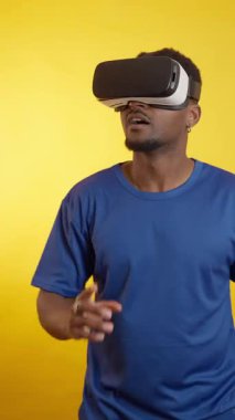 VR kulaklık takan genç bir adam, canlı sarı bir arkaplana karşı, görünmeyen uyarıcılara jest yaparak ve tepki vererek sanal bir çevreyle etkileşime giriyor.