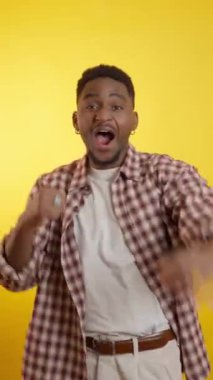 Enerjik genç Afrikalı Amerikalı erkek neşeyle dans ediyor, dinamik bir şekilde sarı arka planda dans ediyor.