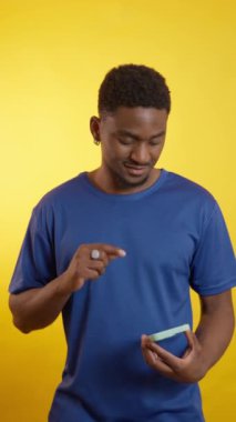 Sarı arka planda izole edilmiş yeşil ekranlı akıllı bir telefonu tutarken çeşitli jestler yapan Afrikalı genç bir Amerikalı.