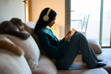 Genç bir bayanın, evcil kedisi eşliğinde kulaklık takıp tablet üzerinde dijital kitaptan keyif aldığı rahat bir ev sahnesi.