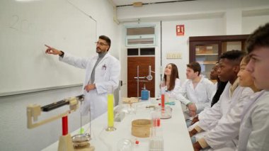 Kimya hocası bilimsel prensipleri gösteriyor, laboratuvar önlüğü giyiyor ve modern laboratuvar ortamında öğrencilerin etrafındaki karmaşık tepkileri açıklıyor.