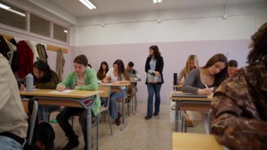 Eğitmen, bir sınıf oturumu sırasında masalarda çalışan öğrencilere odaklanarak, bir akademik enstitüde öğrenim ortamını teşvik eder