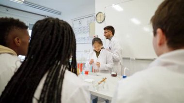 Öğrenciler, bir eğitmenin rehberliğinde aktif olarak bir kimya deneyine katılıyorlar. Sıvıları karıştırmaya ve laboratuvar tablosu ve ekipmanlarındaki tepkileri analiz etmeye odaklanırlar.