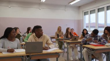 Sınıftaki bir öğretmeni dikkatle dinleyen çeşitli öğrenci grupları, öğrenme ve işbirliği için dizüstü bilgisayarlar ve defterler kullanıyorlar.