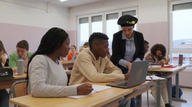 Eğitmen, akademik bir ortamda işbirlikçi öğrenmenin altını çizerek, bir sınıf dersi sırasında laptopta çalışan ilgili öğrencilere yardımcı oluyor.