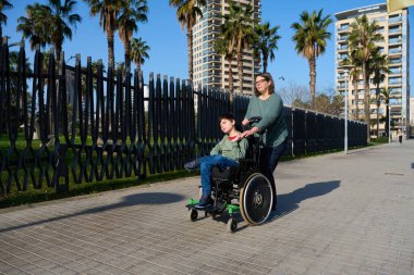 Anne oğluna engelli olarak yardım ediyor, tekerlekli sandalyesini parkın yanındaki kaldırımlı yolda itiyor, erişilebilirliği ve dahil edilmeyi teşvik ediyor.