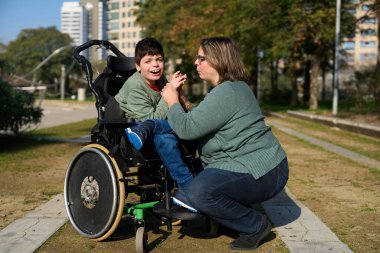 Serebral felçli mutlu çocuk parkta annesiyle el ele oturarak güneşli bir günün tadını çıkarıyor.