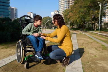 Anne, tekerlekli sandalyede oğlunun yanında çömelmiş, onu parkta teselli ediyor, sevgi ve destek gösteriyor.