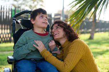 Mutlu anne sevgiyle oğlunu kucaklıyor tekerlekli sandalyede oturuyor, güneşli bir günün tadını çıkarıyor.