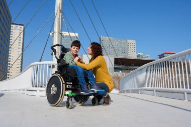 Sevgi dolu bir anne, şehirde bir köprüde ellerini tutarken oğlunu tekerlekli sandalyede rahatlatıyor.