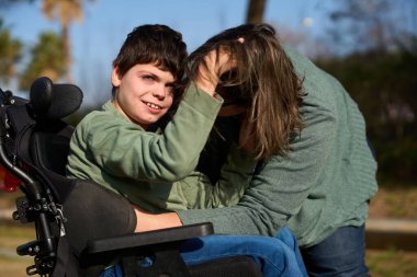 Parkta tekerlekli sandalyesinde güneşli bir günün tadını çıkarırken annesinin saçıyla oynayan serebral felçli çocuk.