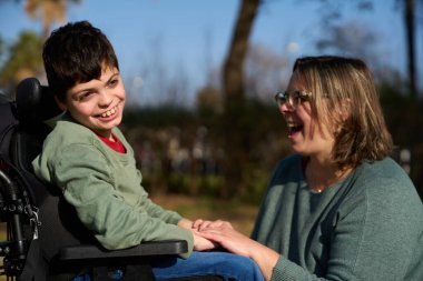 Parkta tekerlekli sandalyede annesiyle el ele tutuşurken gülümseyen mutlu çocuk güneşli bir günün tadını çıkarıyor.