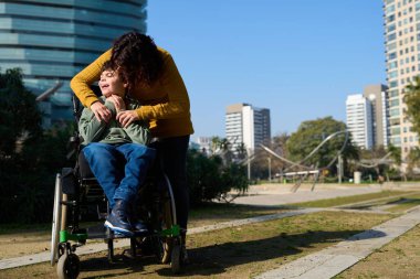Anne engelli oğlunu kucaklıyor, huzurlu bir şehir parkında ona sevgi ve destek sağlıyor.