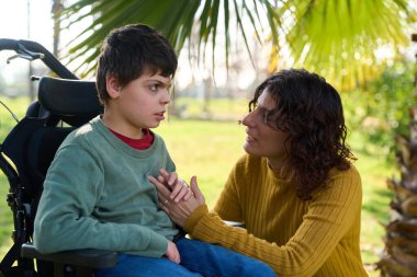 Bakıcı anne, tekerlekli sandalyede oturan oğlunu şefkatle teselli ediyor. Ona huzur parkında güven ve destek veriyor.