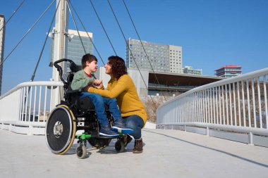 İlgili bir anne, tekerlekli sandalye kullanan genç oğluyla iletişim kurar, modern bir şehir ortamında yaya köprüsünde hassas bir anı paylaşır.