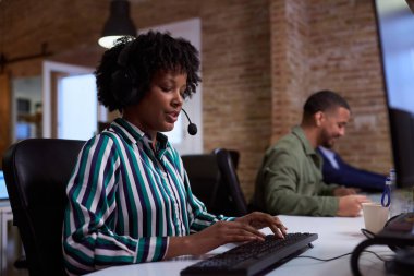 Müşteri desteği sağlayan, klavyeye yazan ve yoğun bir ofis ortamında kulaklığa konuşan odaklanmış bir çalışan.