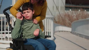 Sıcak kalpli anne gülümseyen engelli çocuğu desteklerken açık havada güneşli bir anın tadını çıkarıyor, şehir köprüsü yürüyüşü sırasında gerçek mutluluğu paylaşıyor.