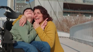Şefkatli bir anne tekerlekli sandalyeye bağlı oğlunu kucaklıyor, şehir köprüsünde samimi bir anda derin bir bağ kuruyor, koşulsuz sevgi ve desteği somutlaştırıyor.