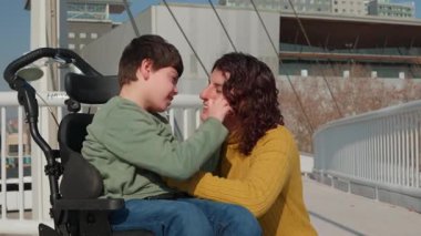 Engelli çocuk, neşeli bir şekilde şehir köprüsünde destekleyici bir anneyle oynuyor, güneşli bir günde iç açıcı bir bağ ve mutluluk anını paylaşıyor.