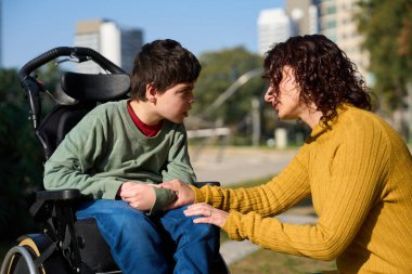 Umursayan bir anne, oğlunu tekerlekli sandalyede destekliyor güneşli bir parkta sohbet ediyor.