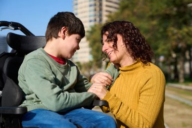 Anne ve çocuk gülümsüyor ve el ele tutuşuyor, parkta güneşli bir günün tadını çıkarıyor, aileyi kutluyor ve engellilerin üstesinden geliyor.
