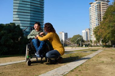 Serebral felçli gülümseyen çocuk parkta güneşli bir günün tadını çıkarıyor, şefkatli annesiyle mutlu anları paylaşıyor.
