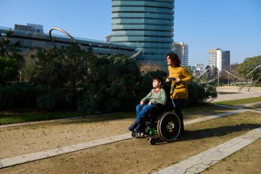 Bakıcı anne oğlunu tekerlekli sandalyeye itiyor, güneşli bir günde şehir parkında eğleniyor.